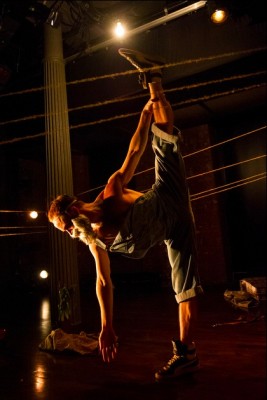 A golden hue outlines a dancer on a stage. Their left leg extends upward, creating a long, vertical shape, juxtaposed by lines of string tied from one side of the set to another. Photo by Ian W. Douglas.