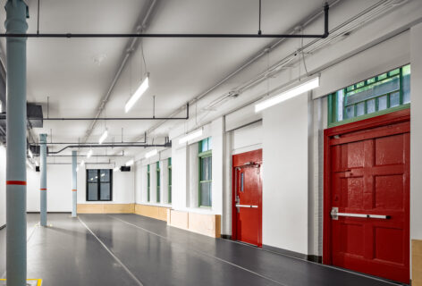 A dance studio with a grey marley floor, windows, and red doors. There are three grey columns distributed through two thirds of the space. Photo by Alexander Severin.