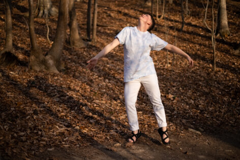 Londs Reuter dancing in a natural setting with fall leaves and trees in the back and foregrounds. She wears all white and stand extending her arms away from her torso. Her head looks upward and diagonally back. Photo courtesy of the artist.
