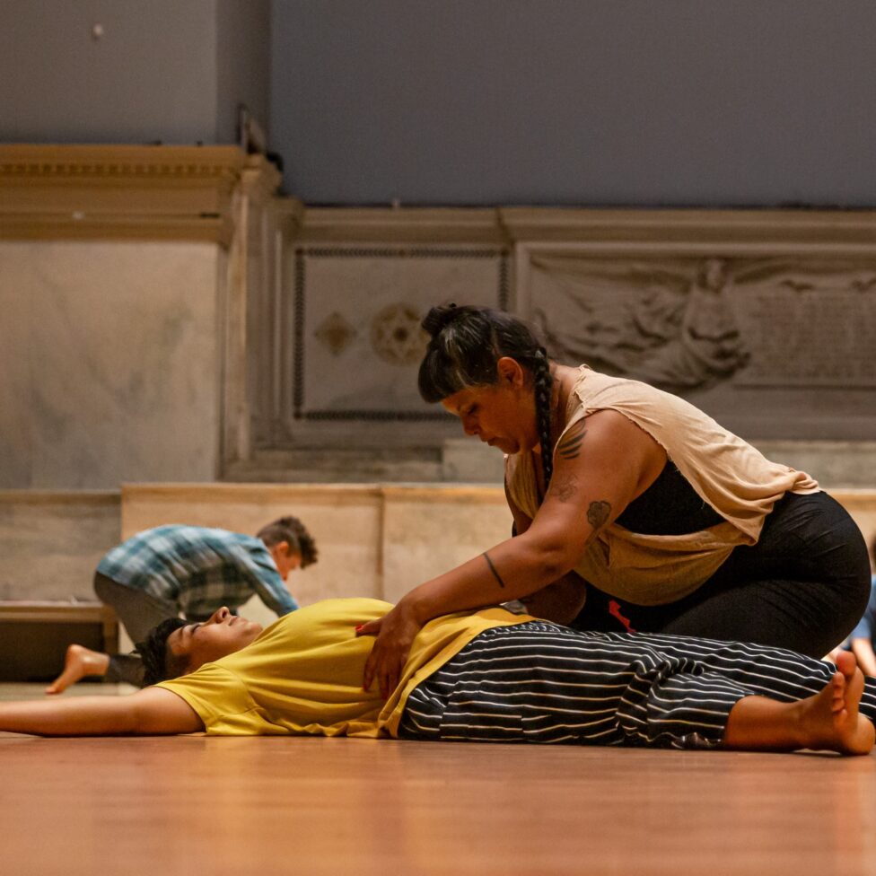 Two dancers at level floor during Sara Shelton Mann's class. One is lying on their back with arms and legs extended out while the other is kneeling next to them with hands gently touching the pelvis of the dancer at rest. Behind them another dancer can be seen doing the same holding gesture. The Judson Memorial Church altar serves as background. Photo by Rachel Keane.