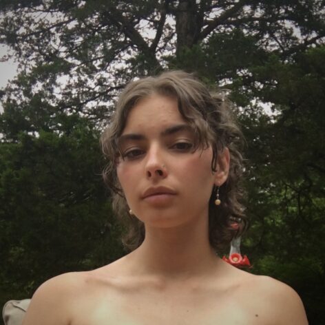 A person with light olive skin and brown curly hair which is partially pulled back. They are wearing earrings, a nose ring, and are backed by a large evergreen tree. Photo by Charline.
