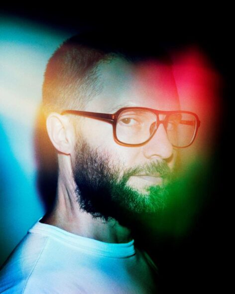 A profile shot of Barnett Cohen. He is wearing black frame glasses and a white shirt. Red, yellow, and green light reflections cover his face. photo courtesy of the artist.