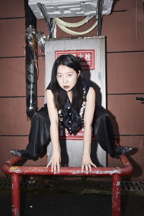 image of Yao Qingmei balancing in a wide squat on a railing with their hands reaching down on a pole. They are in all black with floral patterned shoes and a vest. Behind them, is a red panel wall, they look directly to the camera.