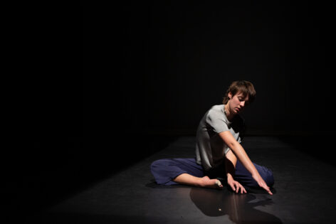 Campbell sits in fourth position on a stage surrounded by blackness, wearing a glittery silver short sleeve, and dark blue open leg pants. Their visible big toe is wrapped in white tape. They have white skin, pixie-short hair, that is dark and straight, and their right arm is extended across their torso as they gaze at the ground past their fingertips. Photo by Julia Dissic.