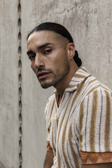 Frederick Quiñones poses for a portrait. He has dark hair pulled back into a low bun. He wears a white and orange striped button up top. He is looking into the camera. Photo by Elisabeth Toll.