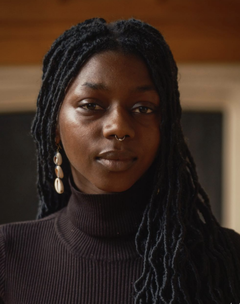 Yacine Fall looks into the camera with a calm expression. She wears a dark turtle neck top and wears her hair in locs. On her ear shines a dangly cowrie shell earring. Photo by Arielle Gray.