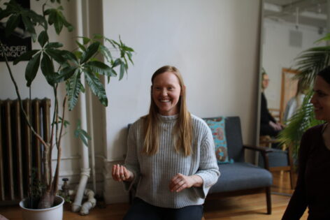 Alice is seated in a chair smiling while teaching in a group class setting, her arms are in a gesture. Photo courtesy of the artist