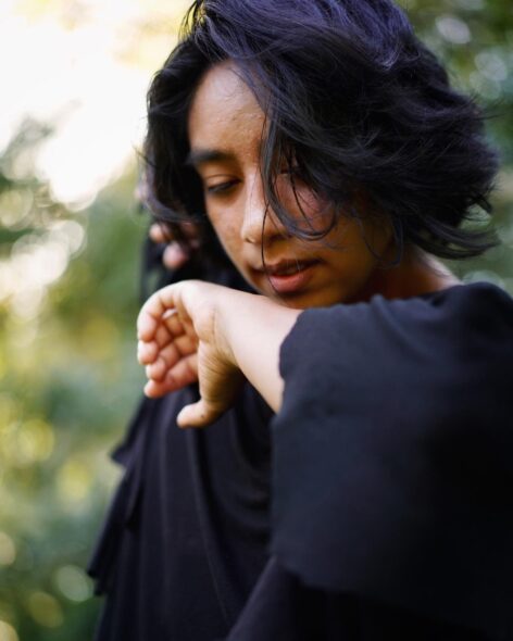 Violeta Tellez right arm bend, hand to chin. She wears a dark top and there is foliage behind her. Photo by Shantel Moses.