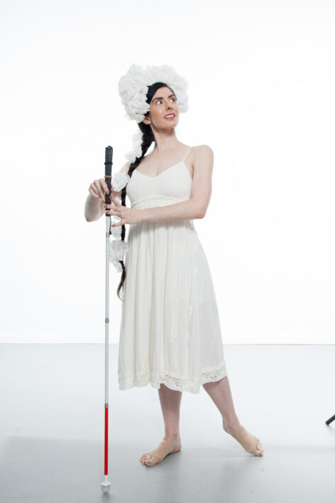 Krishna, a thin, pale skinned woman with long dark hair pulled into a braid, poses on a studio stage. She is wearing a simple white dress with her shoulders exposed and a fluffy white headdress perched on
her forehead and wrapping down her braid. She looks over her left shoulder as she lightly holds a straight cane between her hands and
points her left foot behind her. Photo Credit: Krishna Washburn & Micaela Mamede