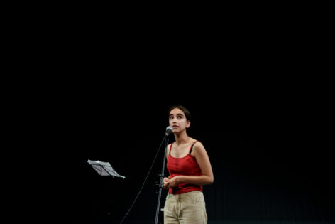 A photo of Leonor standing in front of a microphone looking up. She is wearing a red top and beige corduroy pants. The background is a blackbox. Photo by Joana Linda.
