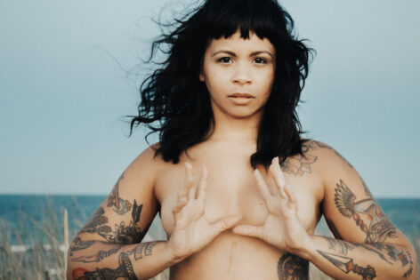 Photo of Marielys Burgos Meléndez a looking into the camera. She is topless with her hands placed in front of her chest, thumbs almost touching and fingers extending pushing the palms outwards. Her arms are tattooed and the ocean in behind her. Photo by Kyle Kesses.
