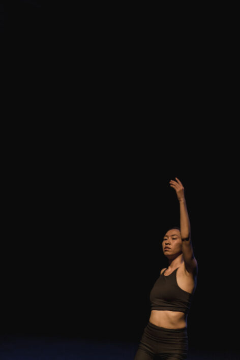 Lai Yi Ohlsen performing. She is standing in darkness with light illuminating her body. She reaches one arm up. Photo by Pratya Jankong.