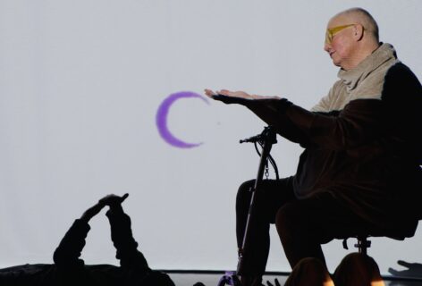 Petra, a white queer disabled cis woman of size sits on her scooter, and reaches out her hand to a video projection of a purple moon. Below, a floor dancer reaches up and touches their own hands. Photo courtesy of the artist.