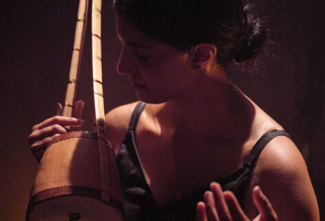 Samita, an Indian-American woman, performs seated, wearing a black dress and her hair pulled back in a bun. She plays an ektaara, a one-stringed instrument, and her eyes are closed in a posture of listening. Photo courtesy of Rachel Topham Photography.