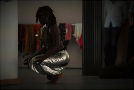 Wearing shimmery pants, a crop top, and a headband, Shantelle Courvoisier Jackson´s hands rest near her knees while squatting. Behind Shantelle, audience members watching. Photo by Scott Shaw.