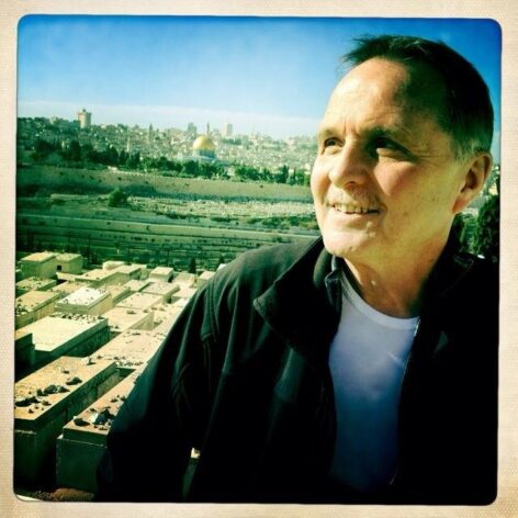Stephen Facey smiles looking beyond the camera. He wears a white top and a black jacket. behind him there is a city scape. Photo courtesy of Stephen Facey.
