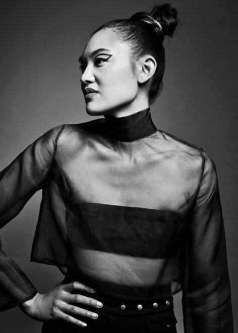 A black and white photo of a Filipinx woman. She looks to the left of the frame. Her right hand is on her hip. She is wearing a sheer turtleneck, through which can be seen a black bandeau top. Her high-waisted pants have 4 tiny buttons across the waist. Her hair is in a bun and her outline-style eyeliner is drawn across her eyelids. Photo credit by Julia Comite.