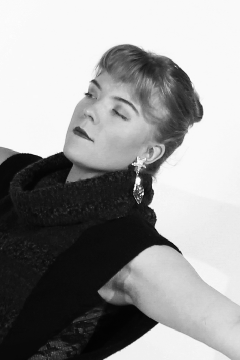 Black and white headshot of Rainey. She is in profile with a serious expression. She has short bangs and her hair is in a bun. She wears a turtle-neck sleeveless sweater and long shiny star earrings. hoto by Photo by Anabelle Scarborough.