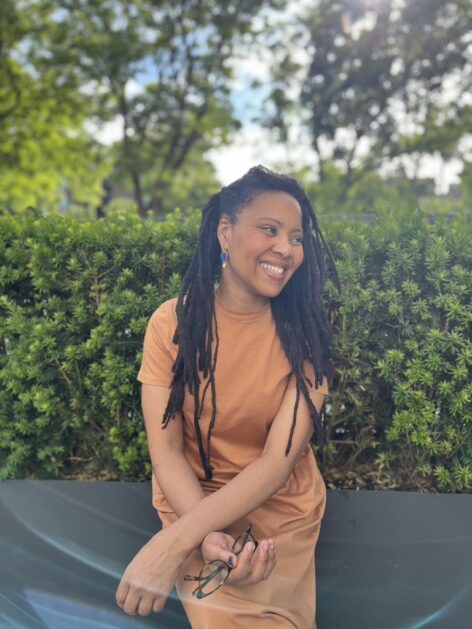Sassy Ma'at smiles while looking away from the camera. She has long dark locs and wears an orange dress. She is sitting in front of greenery. Photo courtesy of the artist.