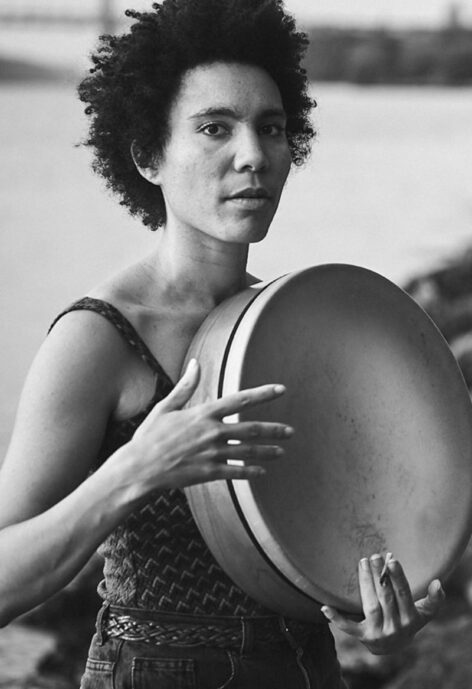 Black and white photo of Anais with a drum, they look toward the camera. Photo by Dar Es Salaam Riser.