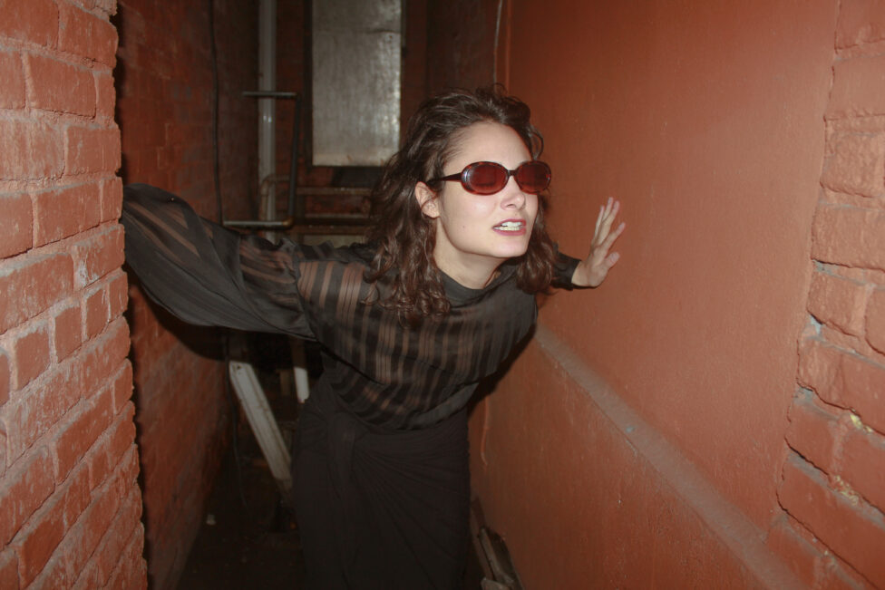 Photo of Anna Witenberg in a brick space, lunging forward, wearing sunglasses.