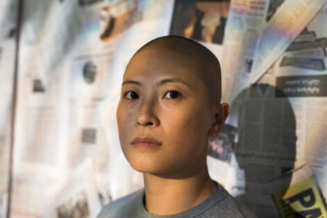 Portrait shot of Annie. She has a shaved head and is looking into the camera with a neutral expression. Photo by An Debie