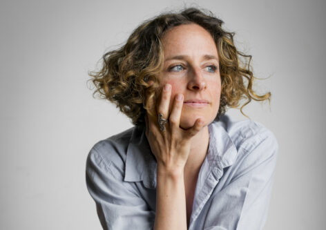 A woman with curly cropped hair, light skin, and blue eyes looks off to the side. Her right hand is up against her face. She's wearing a blue work shirt and is in front of a gray background.