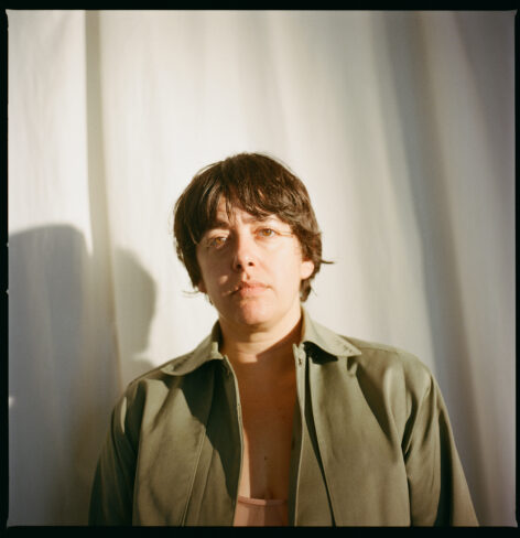 Tess is a white woman with dark brown hair in a short shaggy haircut. In this portrait, she stands in front of a white curtain, squinting slightly with a look of resolve. She wears an olive green trench coat. Bright light falls across one side of her face, illuminating her brown eyes. Photo by Amelia Golden.
