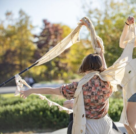 Kiera reaches upward with their right arm, which is wrapped in Sugandha Gupta's sensory textiles made from Kiera's accumulated beige cotton wrist stockinettes (infused with CBD, arnica, and other medicinal herbs after years of use). Kiera's right fingers grip a scarf-like textile held in diagonal tension with Michelle Mantione's crutch, and Kiera's left fingers reach outward to the side toward a lower diagonal chain of stockinettes looped together at the thumb holes as sunlight filters through the fabric. Photo courtesy of the artist.