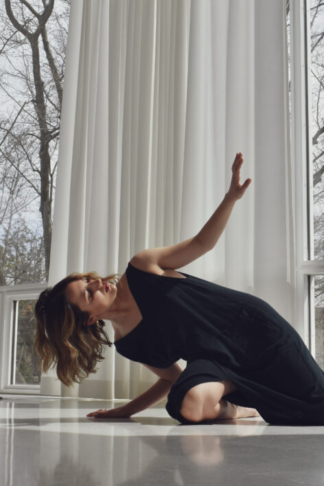 Frances rests on the floor, leaning her weight onto her right arm while looking towards her left hand, positioned above her face. She is wearing a long black dress and her hair is medium-length with Blonde highlights. The room is white and there are barren trees outside the window. Photo by Lee Thomas.