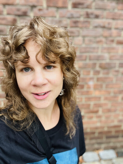 Elinor Tollerz Bratteby smiles into the camera. She wears a dark tshirt with a blue design in the front. He hair is dirty blonde and curly. She stands in front of a brick wall. Photo courtesy of the artist.