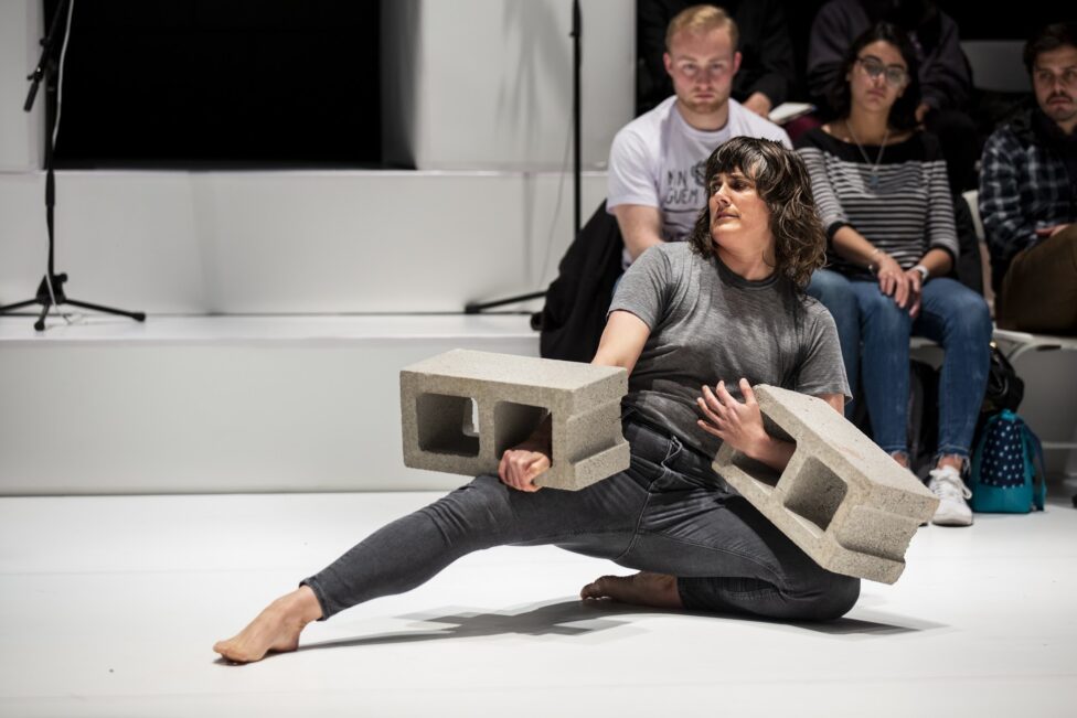 Faye Driscoll performing, wearing a grey outfit and holding two cinderblocks.