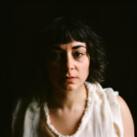Portrait fo Beth Gill. She is lit from the left side of her face. She wears a white top and wears her dark hair loose. She looks into the camera with a neutral face expression. Photo by Maria Baranova.