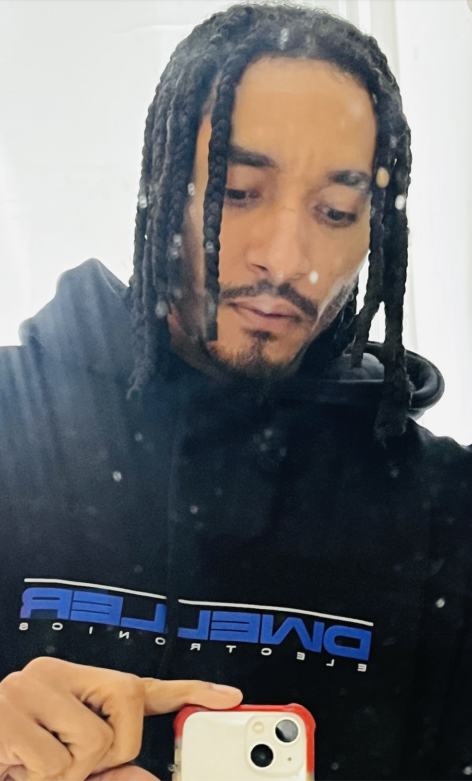 A selfie of Niall Jones. Niall is wearing a black hoodie with blue letters and wears his hair in braids. Niall takes the image with an I-phone pointed at his reflection. Photo by Niall Jones.