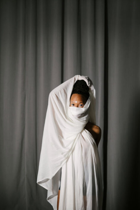 Portrait of Imani Rameses. She wears a long white dress and uses a white fabric to cover the bottom of her face. Her eyes look into the camera. Photo courtesy of the artist.
