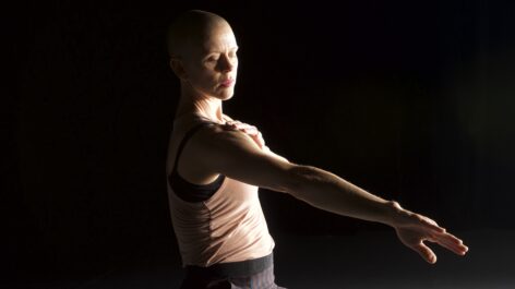 A white woman with a shaved head, wearing a beige tank top. the background is black and she is lit by side light. her focus in downcast, looking at an extended arm and the other hand is gently touching the shoulder. Photo by Jim Coleman