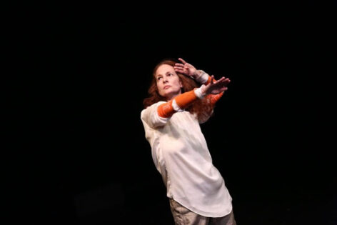 Jodie is wearing light taupe pants, a silky white blouse with bright orange socks as sleeves with a bit of the blouse poking thru the cut out heel of the sock. Her left hand up against my left temple, elbow sharply bent, my right arm straight, extended forward. Her eyes are gazing up to the right high diagonal, looking for Charlie Atlas. Photo courtesy of the artist.