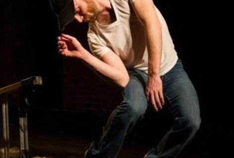 A figure in a white t-shirt and jeans leans in a crouched position, their right hand lightly holding the black baseball cap atop their head. Photo by Ian W. Douglas.