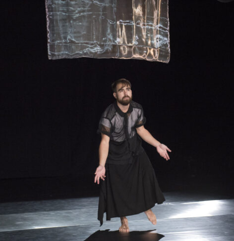 Julian standing under a rectangle set piece, wearing a mesh shirt and black skirt, which his arms out stretched in a performance.