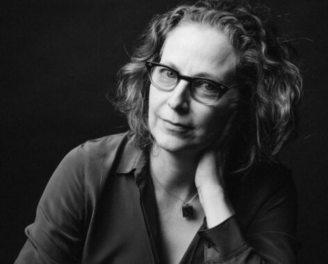 A close-up of a white woman with brown shoulder-length hair and wearing glasses and a teal blue shirt is seated leaning into her left hand which is on her neck. She has a slight smile on her face. Photo by Ebru Yildiz.