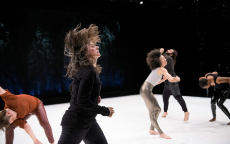 A photo of dancers in motion. In the foregrounded and in profile, the long hair of a dancer whose eyes are closed is suspended above her head.; other dancers behind her are doing individuated movement, seemingly striking or grasping the air. Photo by Ian Douglas.