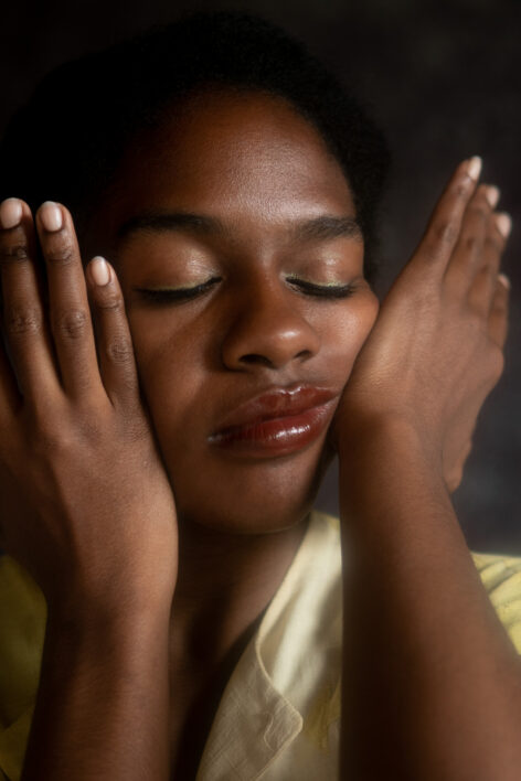 Kayla has her eyes closed and squishes her face in a yellow dress in front of a gray background- imagining a new tomorrow. Photo courtesy of the artist.