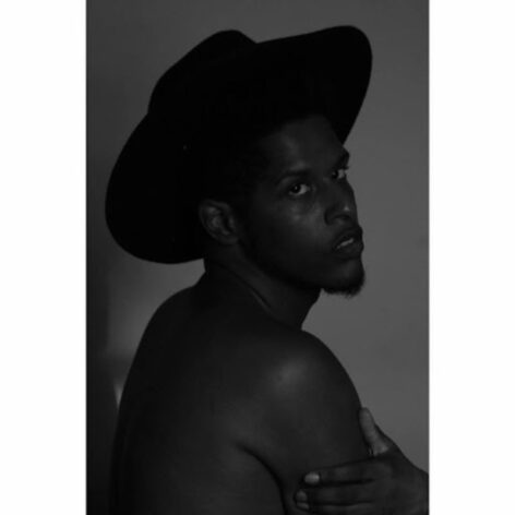 Portrait of Malcolm-x Betts. He is in profile looking over his shoulder towards the camera. He wears a dark fedora hat and is topless. The photo is in black and white.