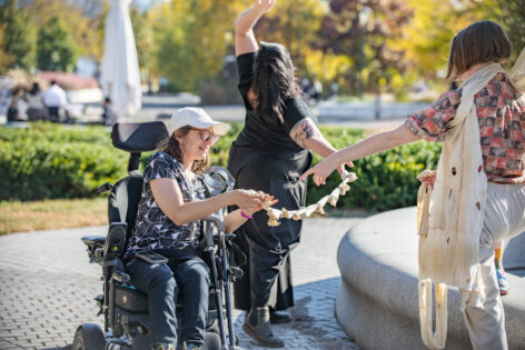 Three dancers wearing pedestrian clothing in an outdoor garden courtyard are posed in a loose triangle formation. Krista holds one end of a chain of connected, beige, cotton wrist stockinettes while reaching upward toward the sun and extending her leg behind her. Michelle holds the other end of the stockinettes chain with both hands while smiling and facing Kiera who wears and holds scarf-like sensory textiles made by Sugandha Gupta out of Kiera’s used wrist stockinettes and points their index finger toward Michelle. Photo courtesy of the artists.