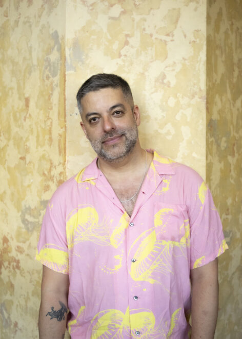 A cis, Latinx man with short dark hair and a greying beard looks directly at the camera with a slight smile. He is wearing a pink shirt with yellow jellyfish print and is photographed against a yellow marbled background. Photo by Chloe Cusimano.