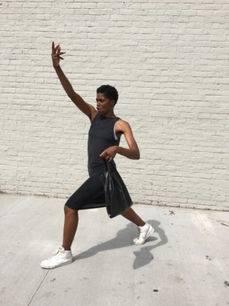 NIC Kay in a black dress and white sneakers stand lunging forward with one arm up, They have a black plastic bag in their hand and mouth open in an o shape.