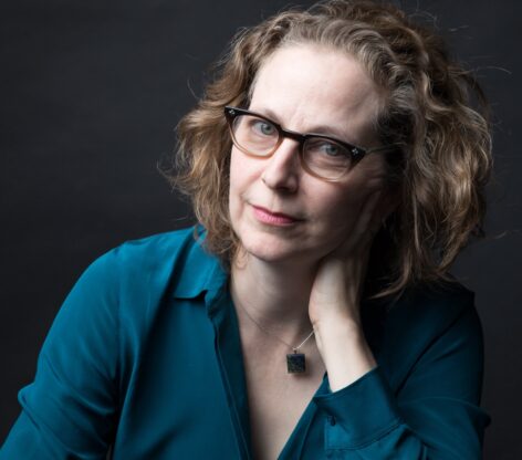 A close-up of a white woman with brown shoulder-length hair and wearing glasses and a teal blue shirt is seated leaning into her left hand which is on her neck. She has a slight smile on her face. Photo by Ebru Yildiz.