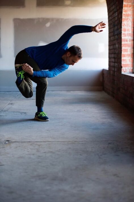 Image of a male dancer in dark green pants, and long sleeve blue shirt. arms are folded in front of him with right arm touching his collarbone. He is leaning backwards in a slight arch.
