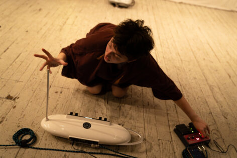 Raychel, a white nonbinary person with short brown hair, kneels on a white floor playing their theremin in a dark brown shirt. Photo by Owen Burnham.