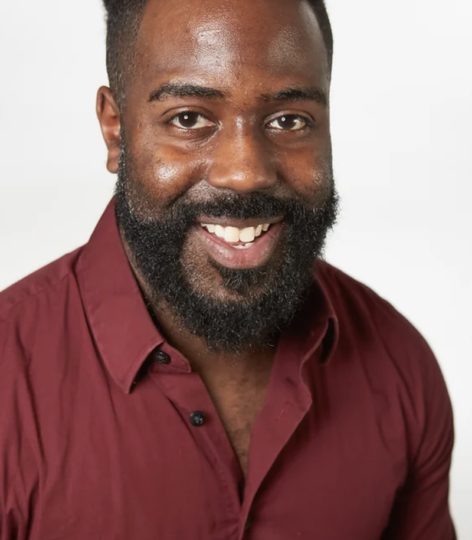 Headshot of Shawn Rawls smiling into the camera. He has a beard and wears a wine colored button up top. Photo courtesy of the artist.